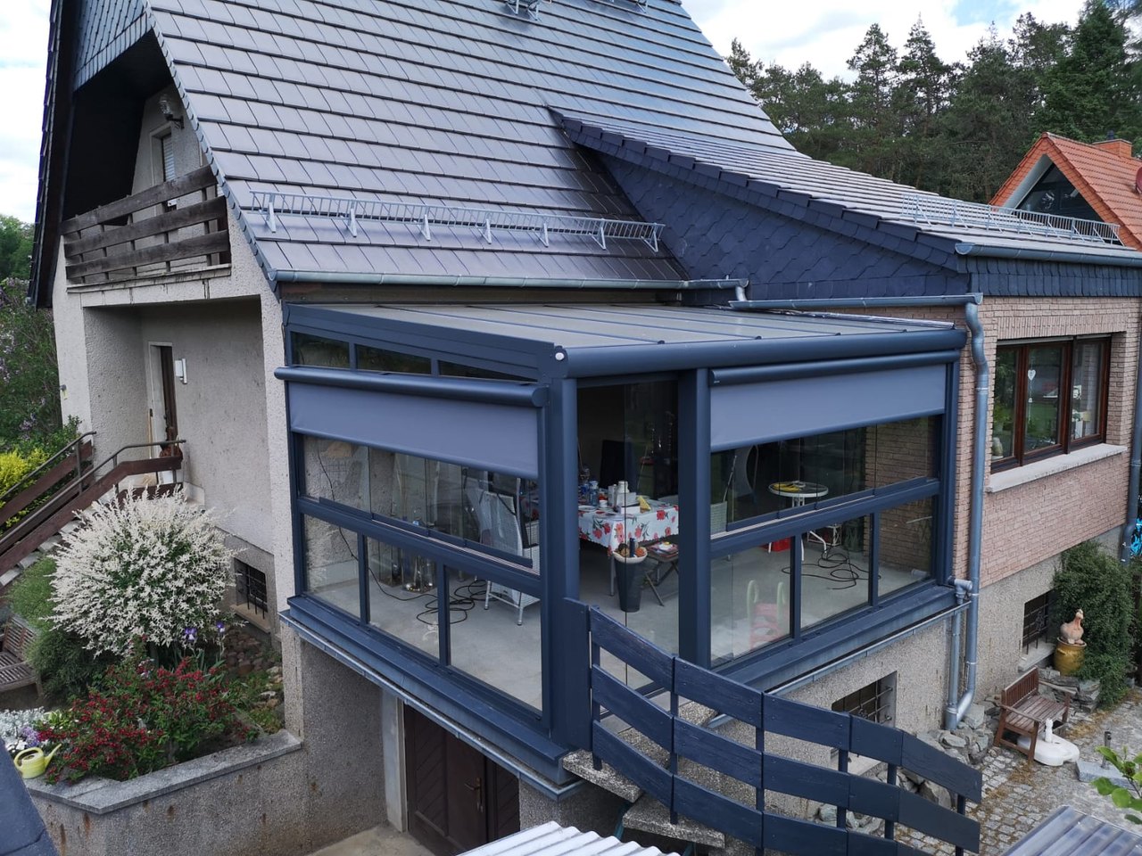 Ein Wintergarten mit grauem Rahmen und Jalousien, der an ein Haus mit Ziegeldach angebaut ist.