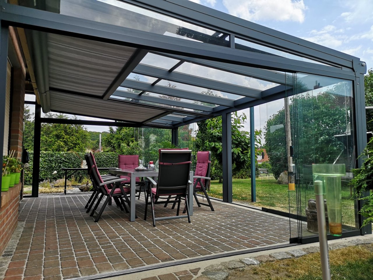 Terrassenüberdachung mit Glasdach und ausziehbarem Sonnenschutz über einem Esstisch mit Stühlen.