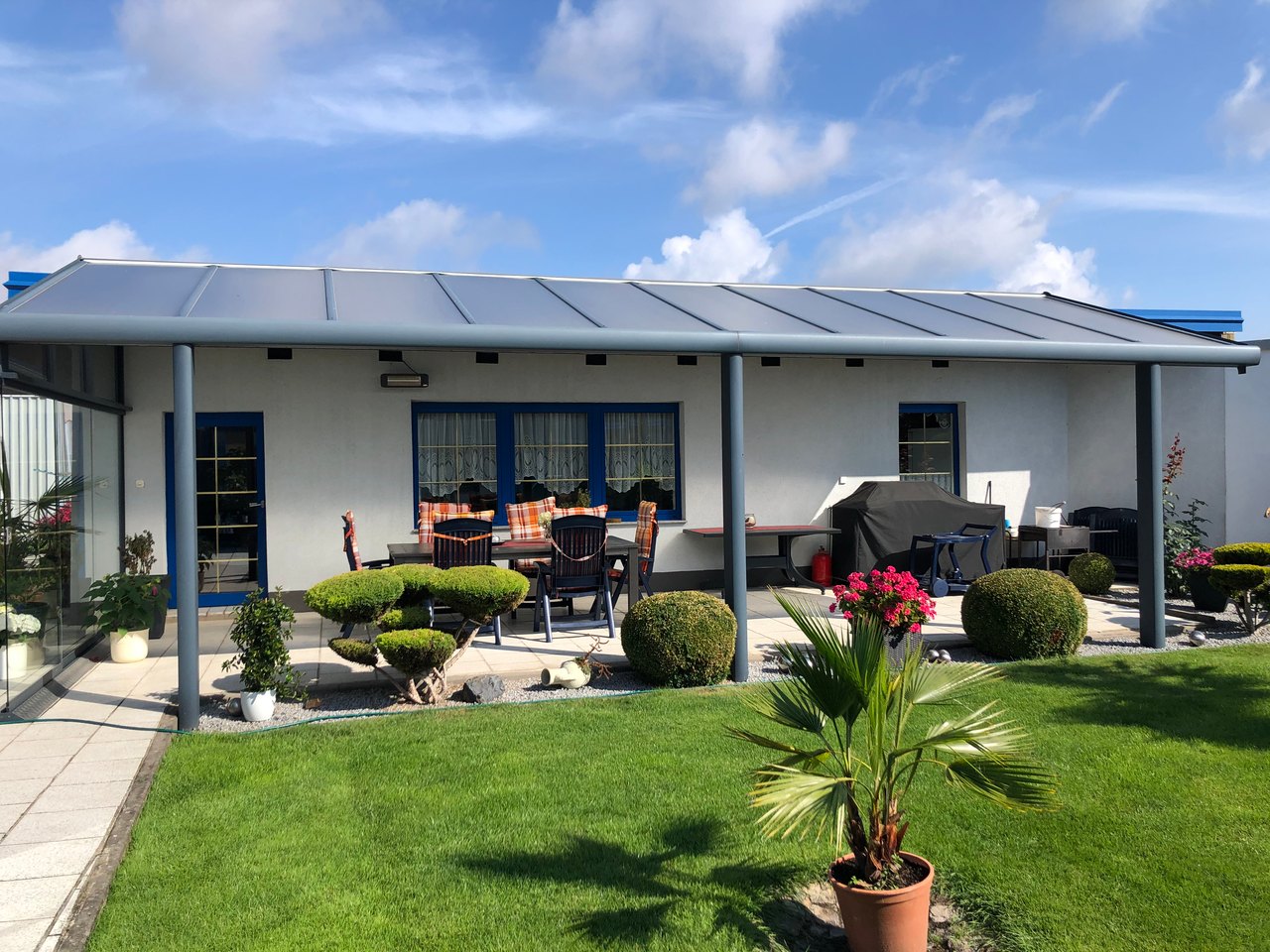 Hinterhof mit überdachter Terrasse, grüner Rasenfläche, Büschen und einem blauen Himmel.