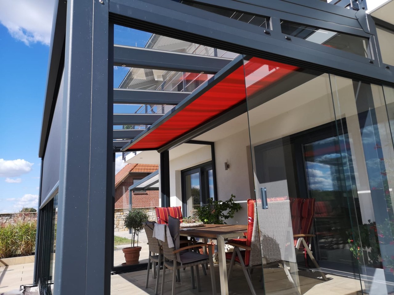 Graue Terrassenüberdachung mit roter Markise und Glastüren, darunter ein Holztisch mit Stühlen.