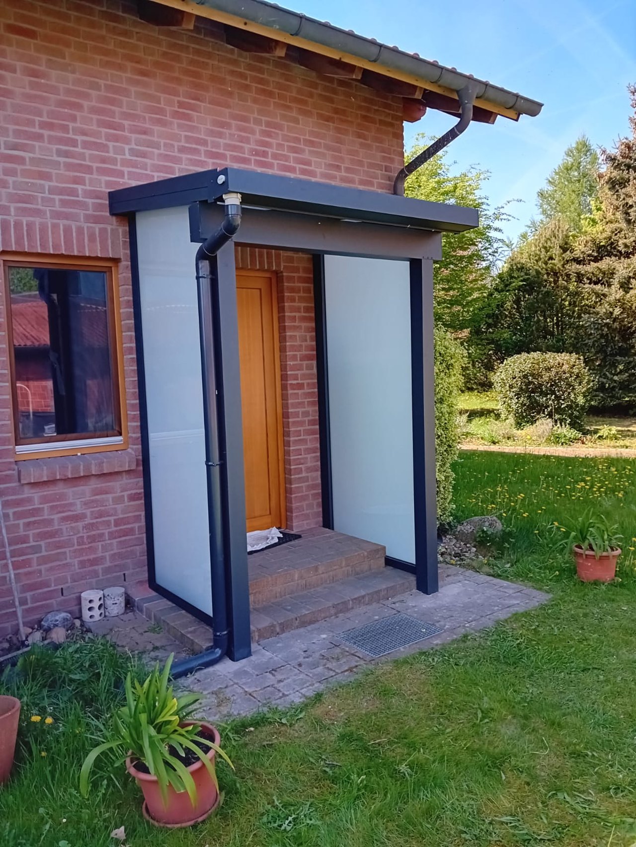 Ein dunkelgraues Vordach mit Milchglas-Seitenwänden schützt eine braune Haustür an einem roten Backsteinhaus.
