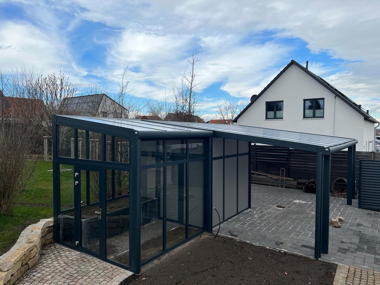 Moderne, anthrazitfarbene Terrassenüberdachung mit seitlichem Gewächshaus und Carport auf gepflastertem Hof.